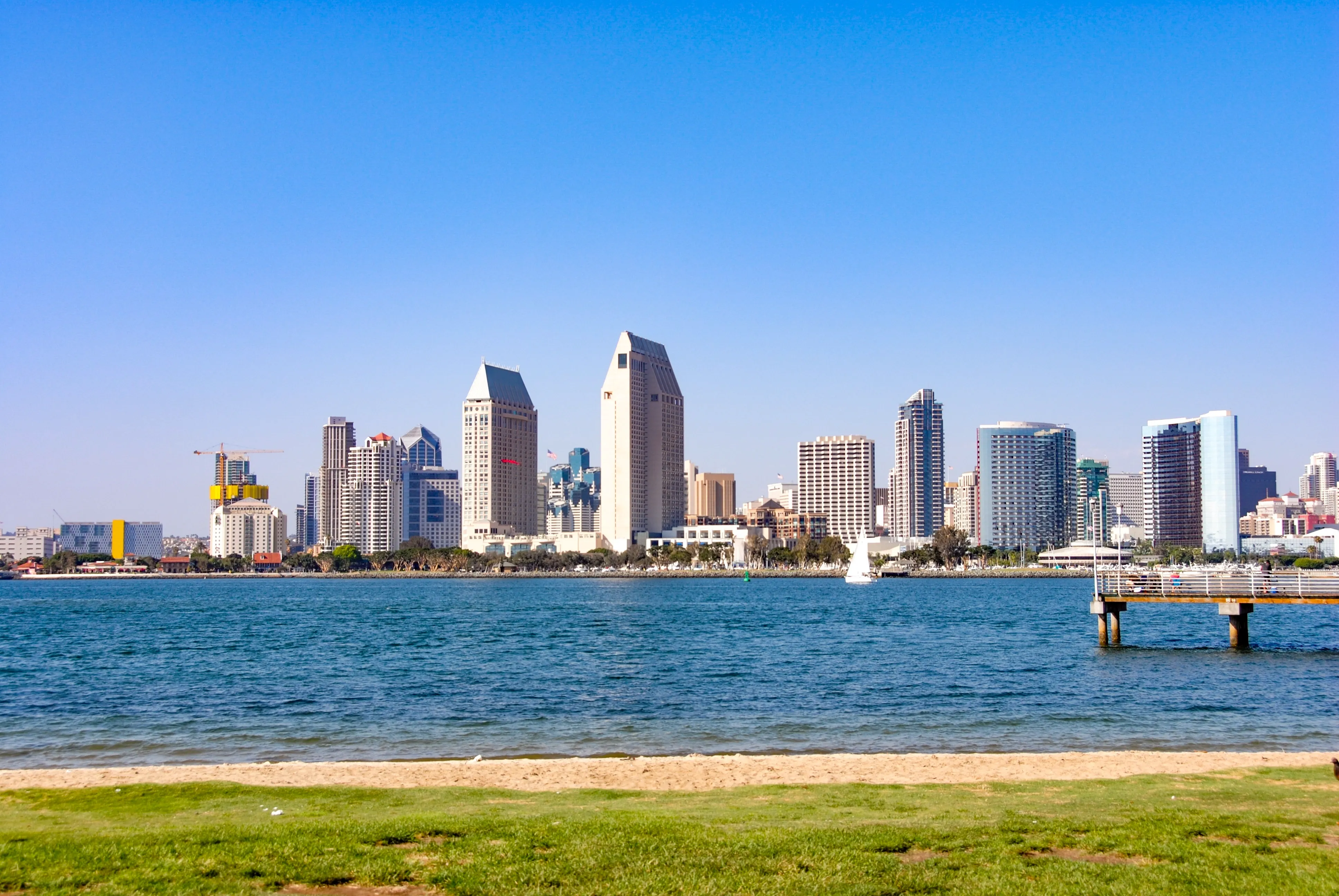 San Diego skyline viewed across blue water, with a grassy shoreline and small pier in the foreground.