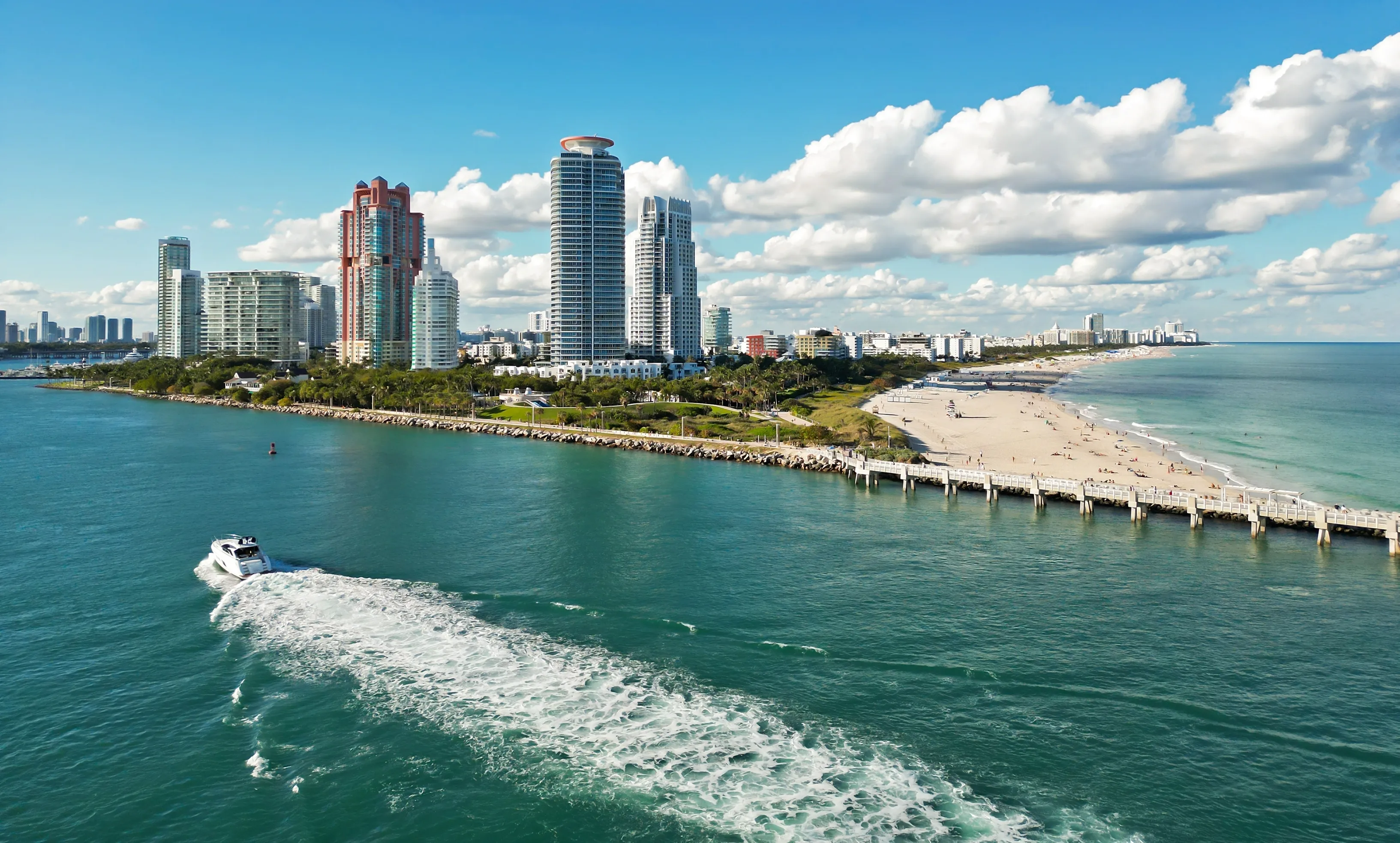 Aerial view of Miami Beach with high-rise buildings, sandy shoreline, turquoise water, and a speedboat leaving a white wake.