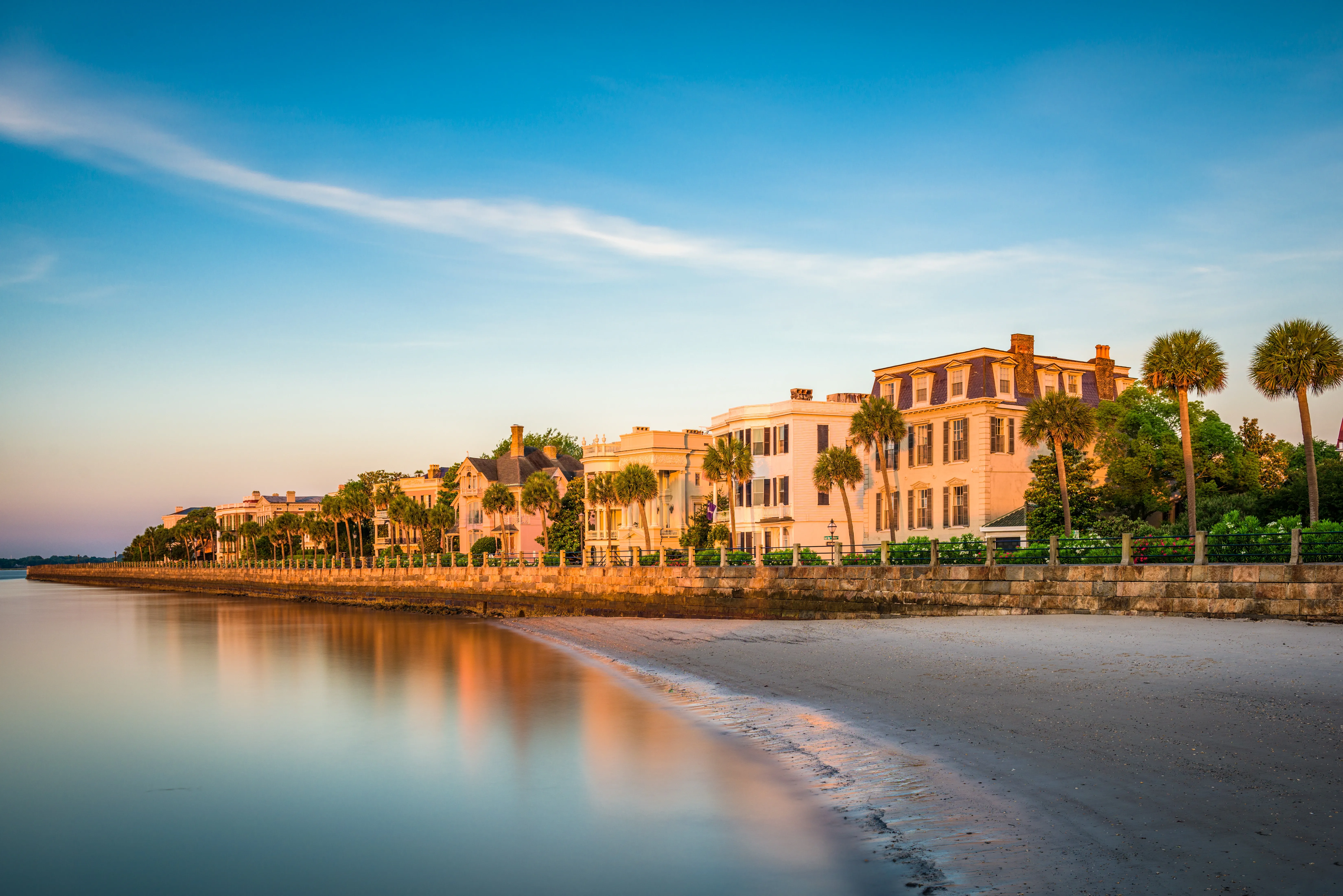 Historic waterfront homes and palm trees line a calm shoreline in Charleston at golden hour.