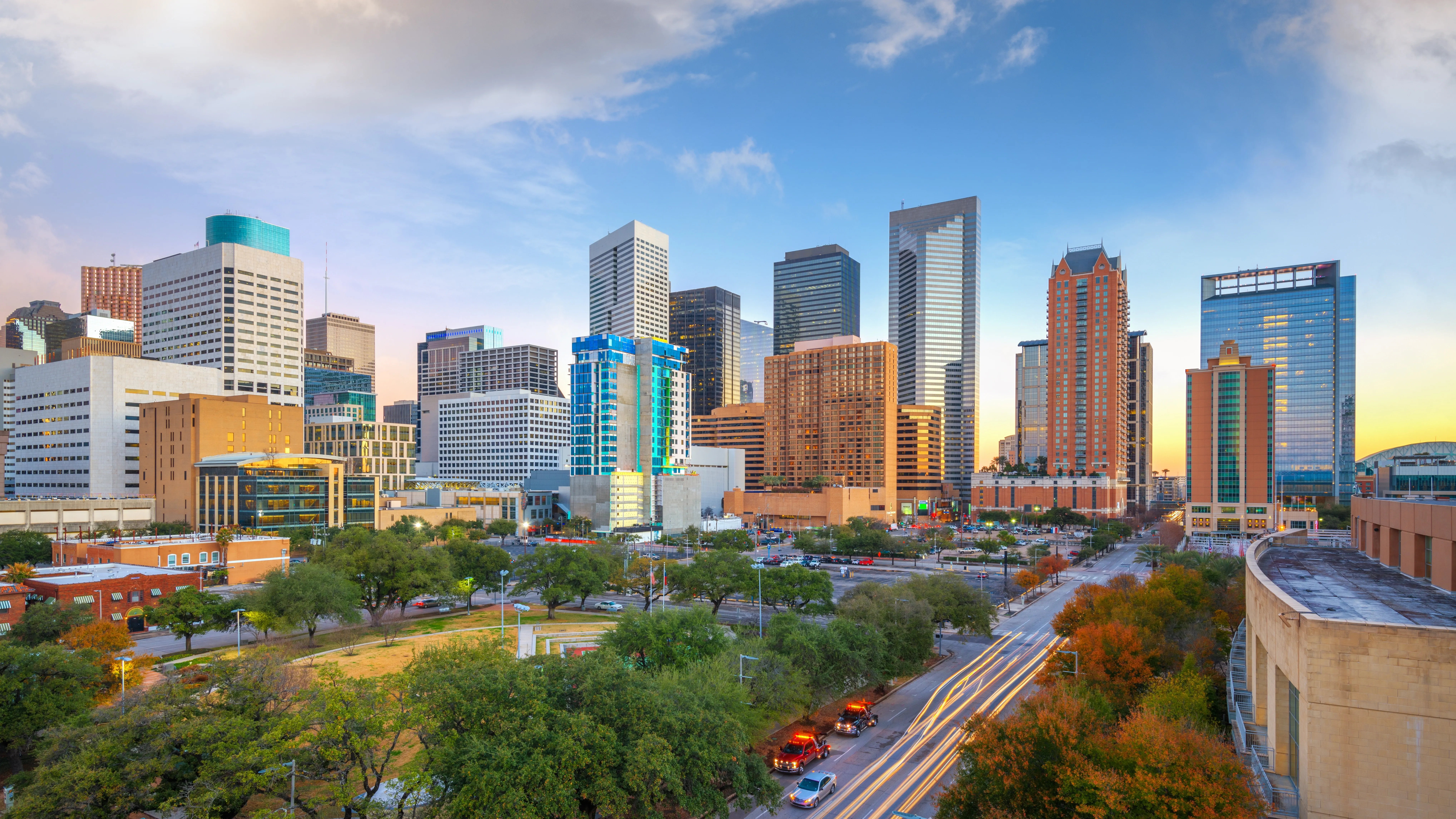 Downtown Houston skyline at dusk with high-rise buildings, city streets, trees, and light trails from traffic.