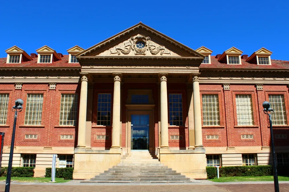 Historic red-brick University of Adelaide building with tall columns, central staircase and clear blue sky.