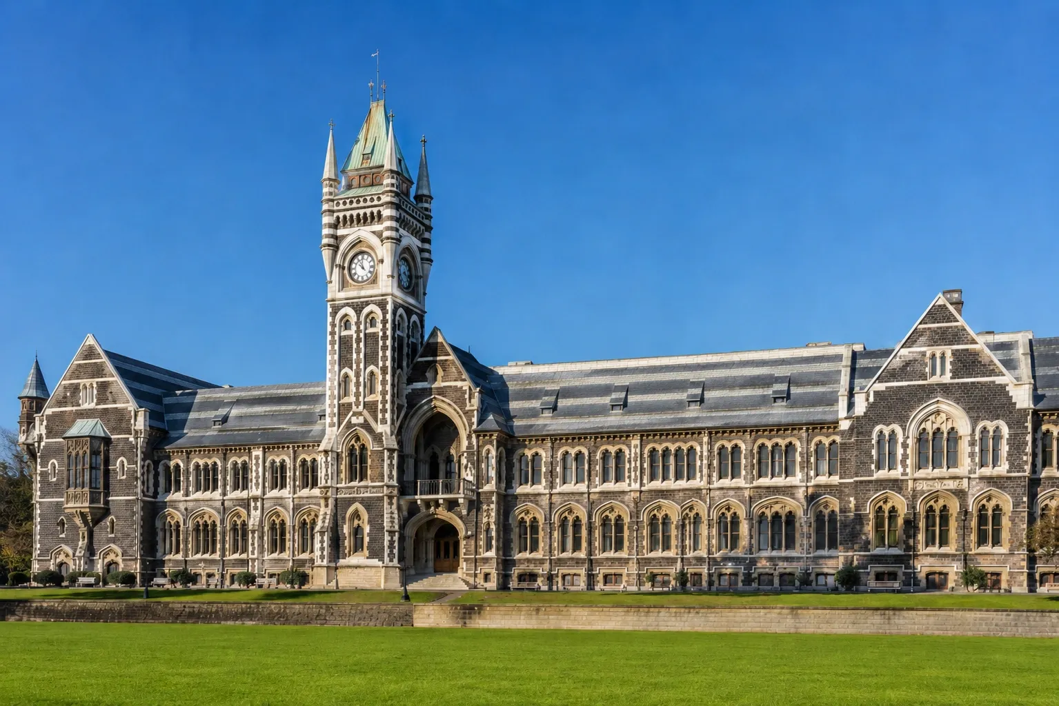 Historic Gothic-style University of Otago building with a clock tower, green lawn and clear blue sky in Dunedin, New Zealand.